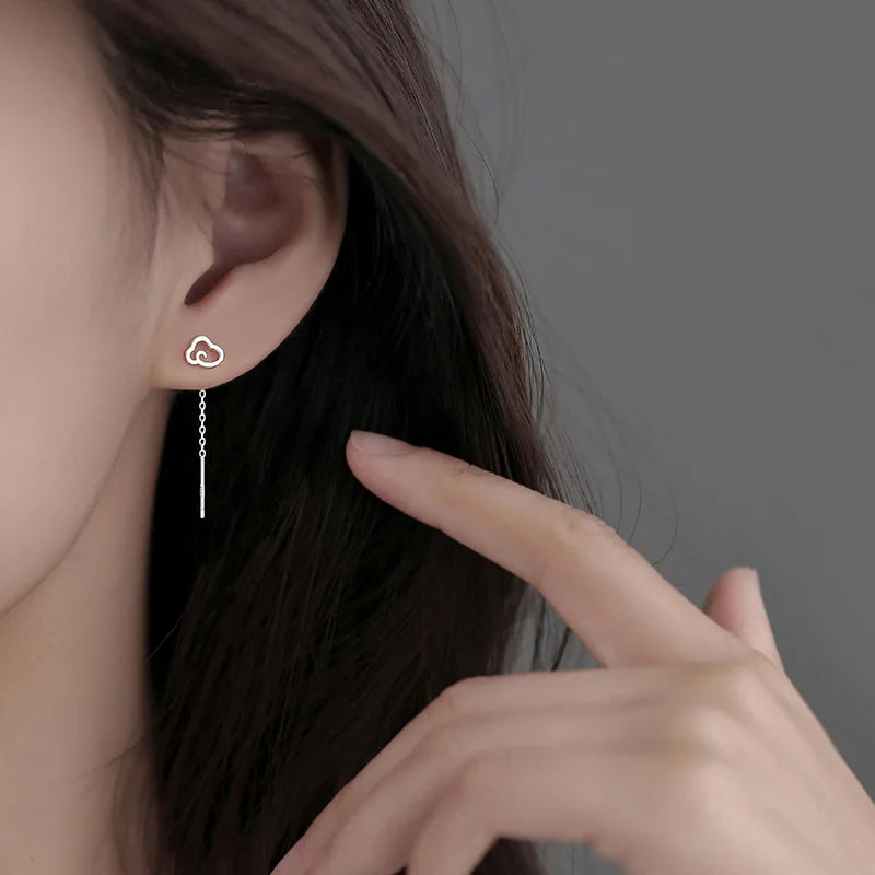 Close-up of a person wearing a silver heart-shaped earring with a thread, against a neutral background.