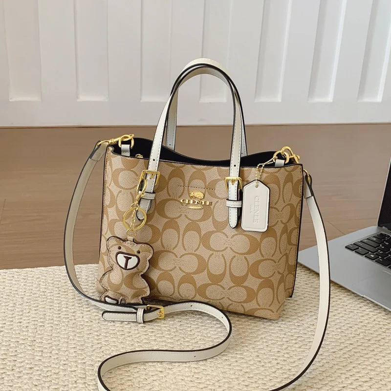Beige handbag with a teddy bear charm on a wooden surface with a laptop in the background