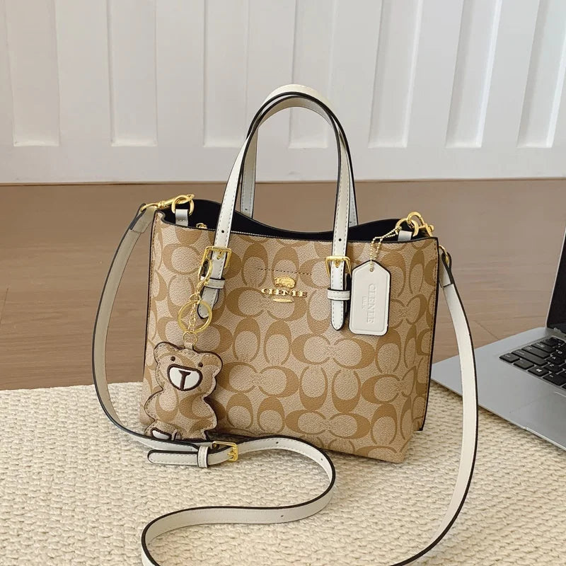 Beige handbag with a teddy bear charm on a wooden surface with a laptop in the background