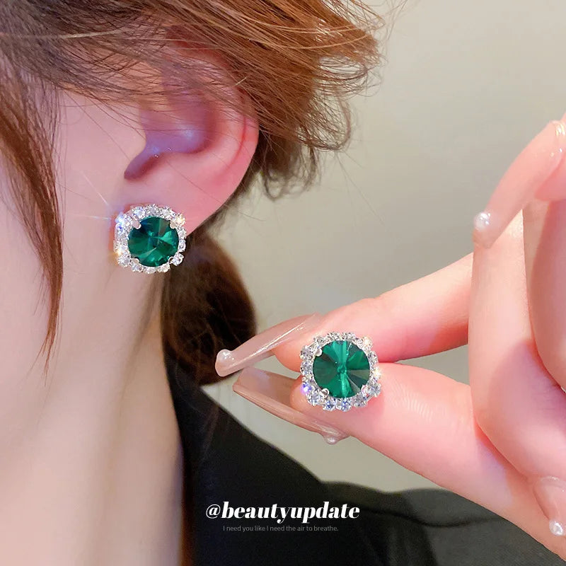 Green gemstone earrings being held near an ear with a neutral background