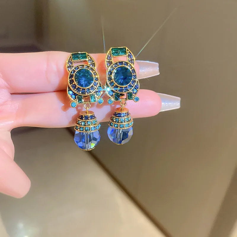 Gold earrings with blue and green gemstones held by a hand against a neutral background