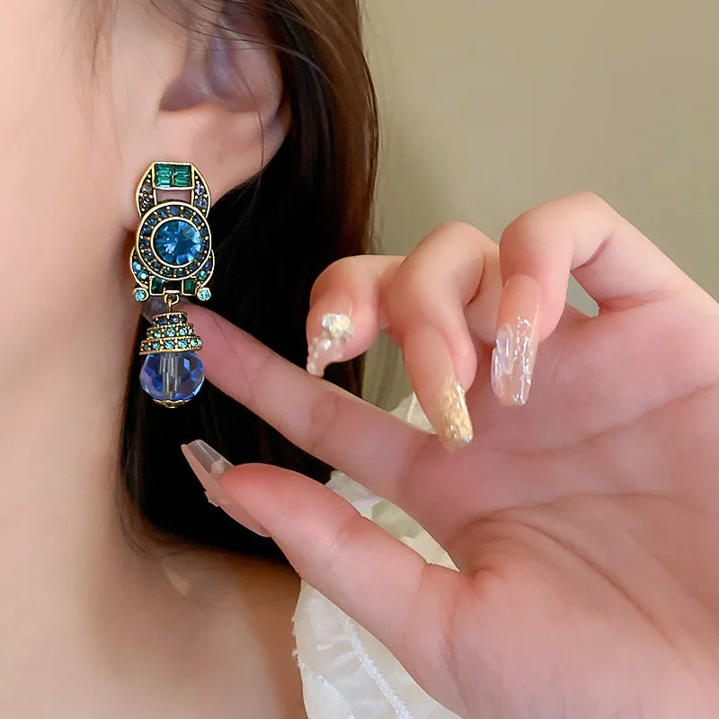 Close-up of a person wearing an ornate earring with a blue gemstone, held by a hand with decorative nails.